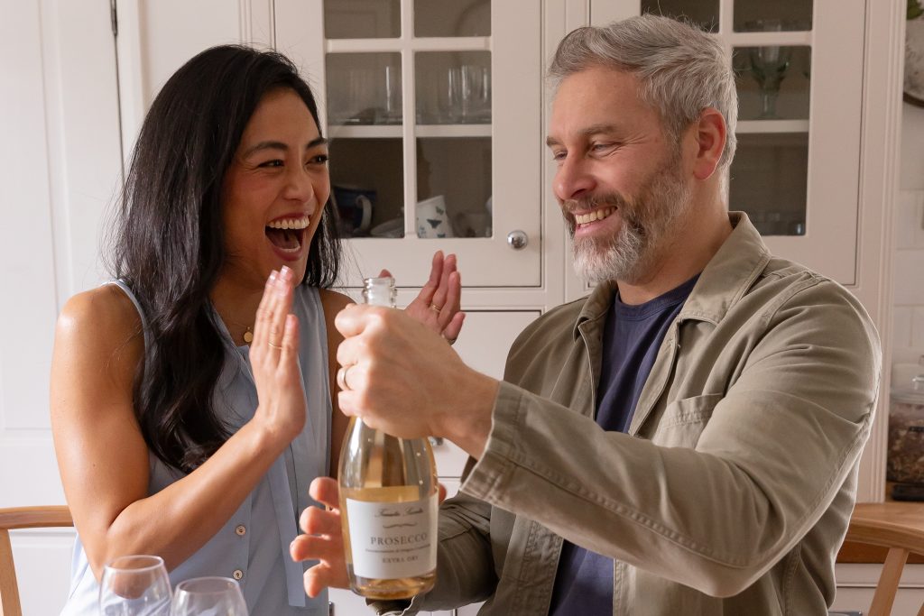 This image shows two people celebrating a significant milestone. They are opening a bottle of prosecco