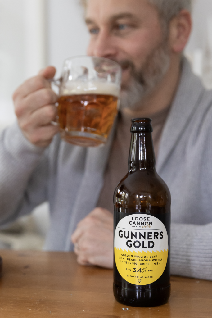 This image shows a man drinking Loose Cannon Brewery's Gunners Gold session Beer. This beer has a yellow label and is included in a gift hamper curated by hampers.com called The Gentleman's Tea Hamper.